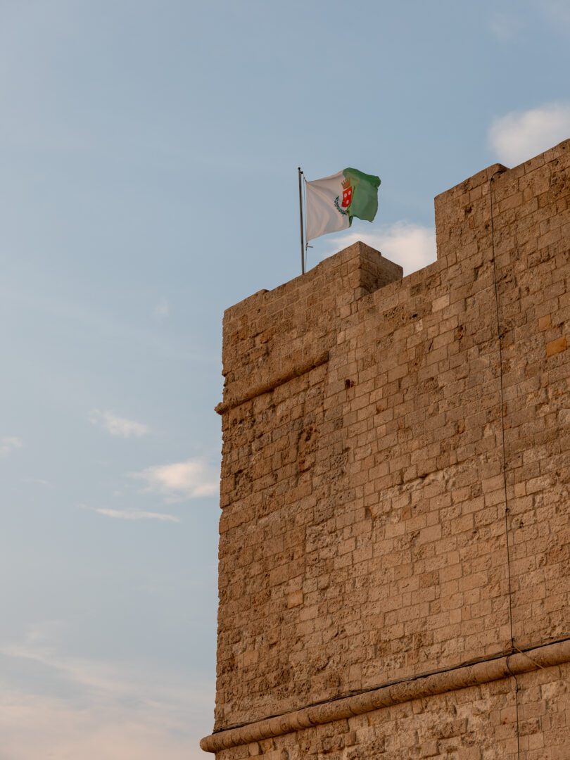 A medieval stone tower of a historic castle in Monopoli, Italy, with the city’s green-and-white flag waving atop against a soft blue sky.