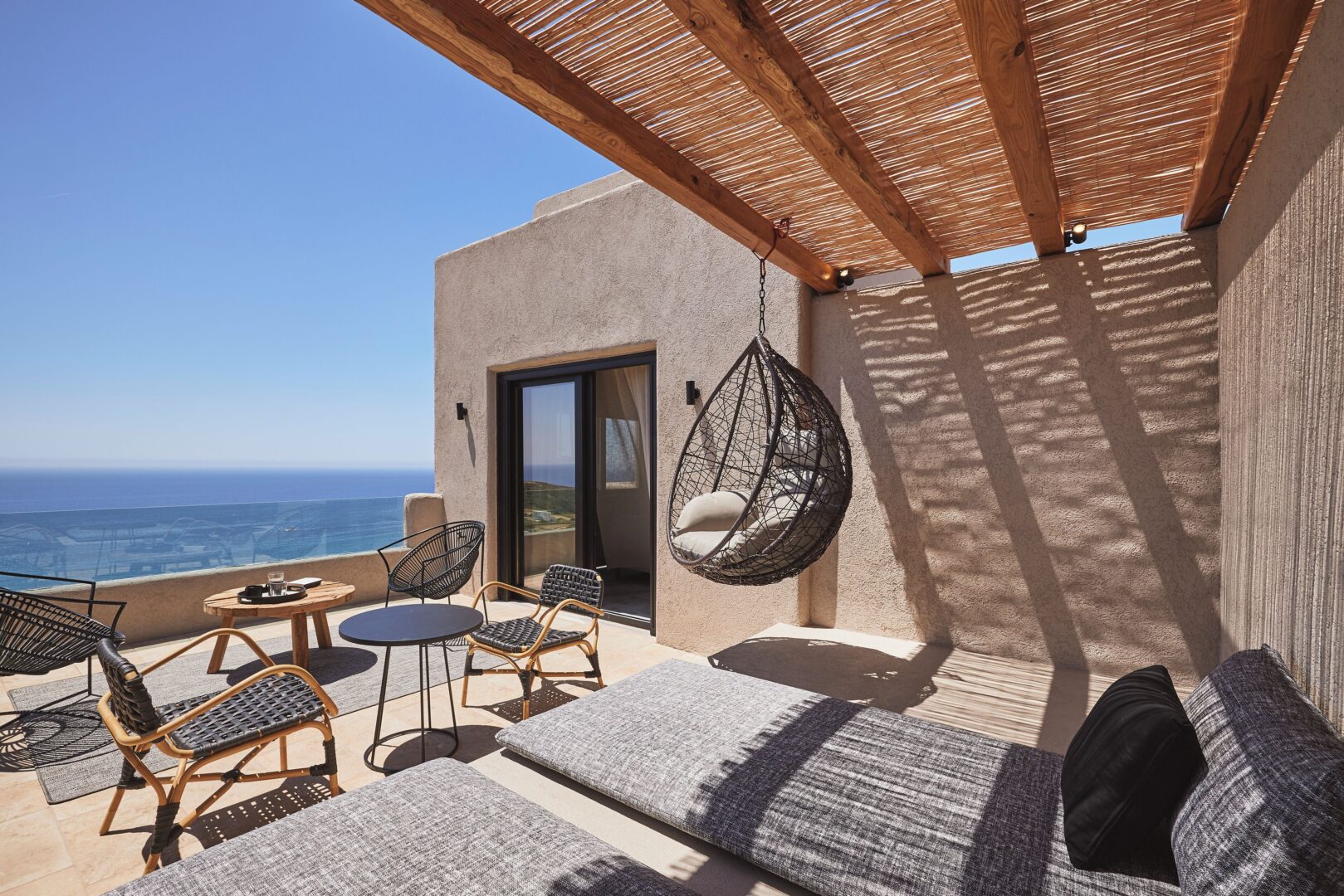 Chic cliffside patio at Noma Milos with hanging chair, sunbeds, and panoramic sea views under a natural wood pergola.