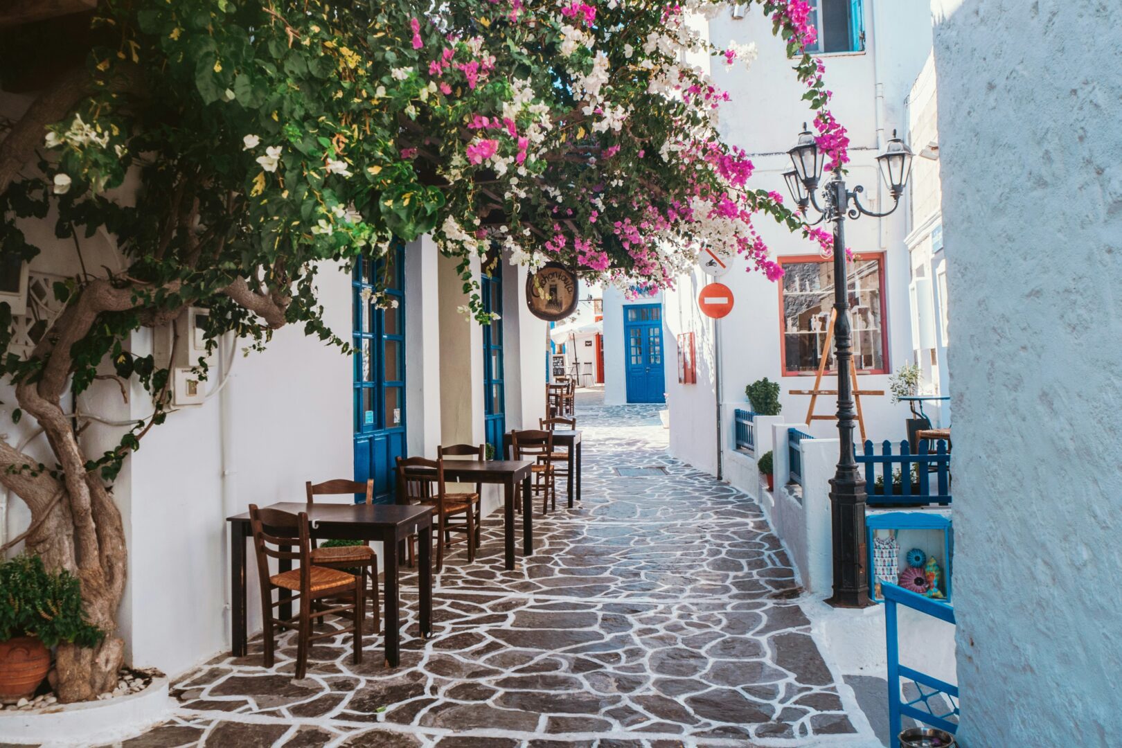 Charming alleyway in Plaka, Milos featuring whitewashed buildings, bright blue doors, and blooming bougainvillea cascading over a café-lined cobblestone path.