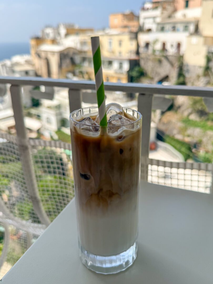 Iced latte with a green-striped straw set against a scenic Fornillo hillside view from the Posides restaurant balcony.