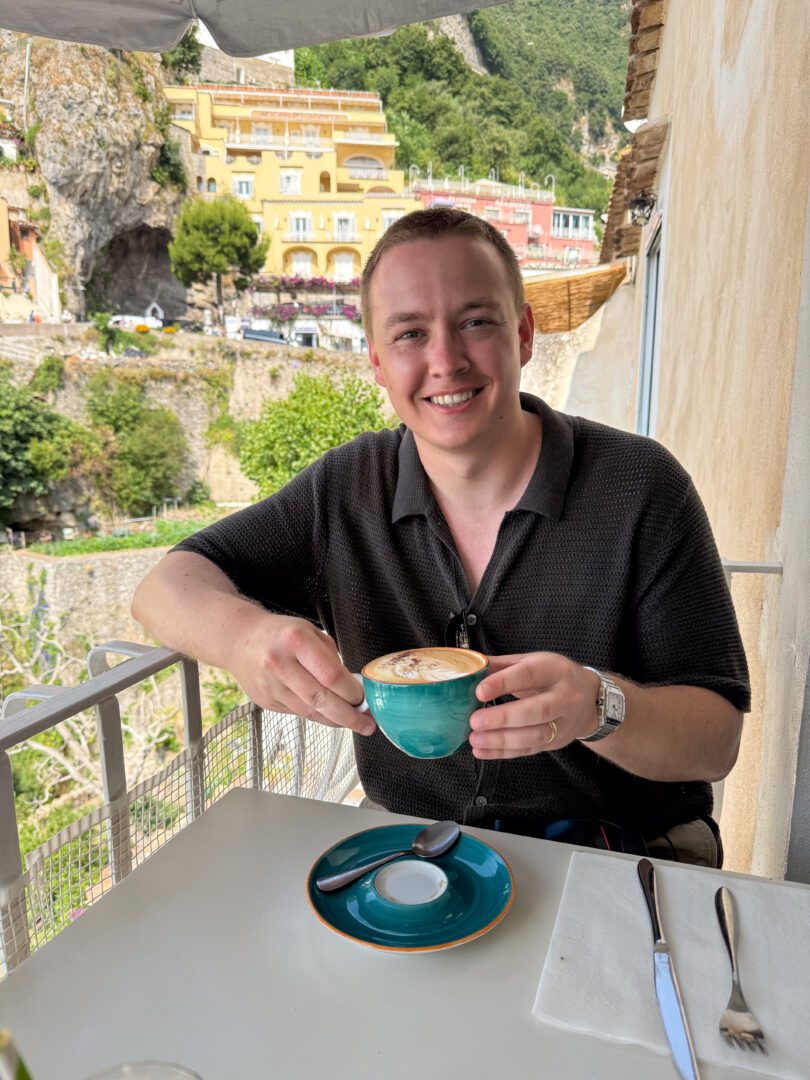 Man enjoying a cappuccino on the balcony of Posides restaurant in Positano, with colorful buildings and cliffs in the background.