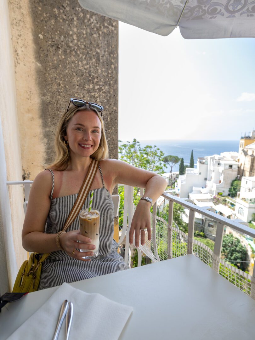 Woman relaxing with an iced coffee on the Posides restaurant balcony, overlooking the sea and rooftops of Positano.