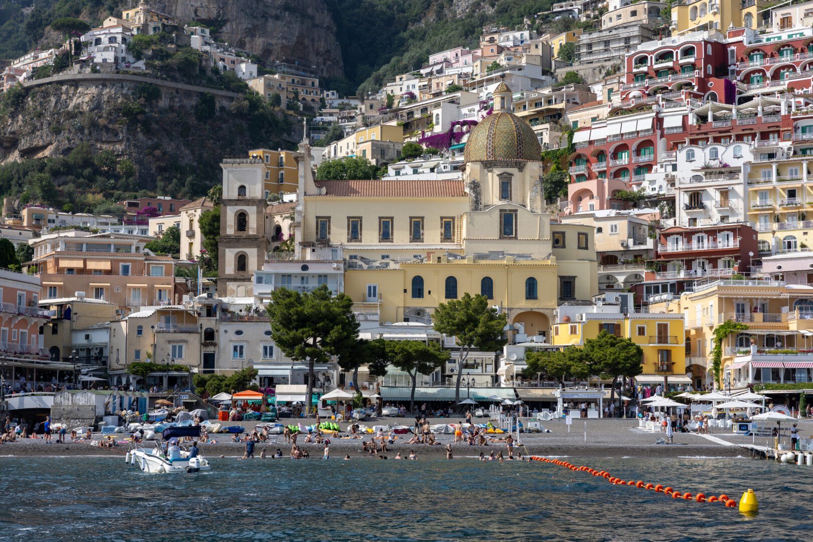 Colorful hillside buildings frame the iconic tiled dome of Santa Maria Assunta as seen from the water, with beachgoers and boats along Spiaggia Grande.