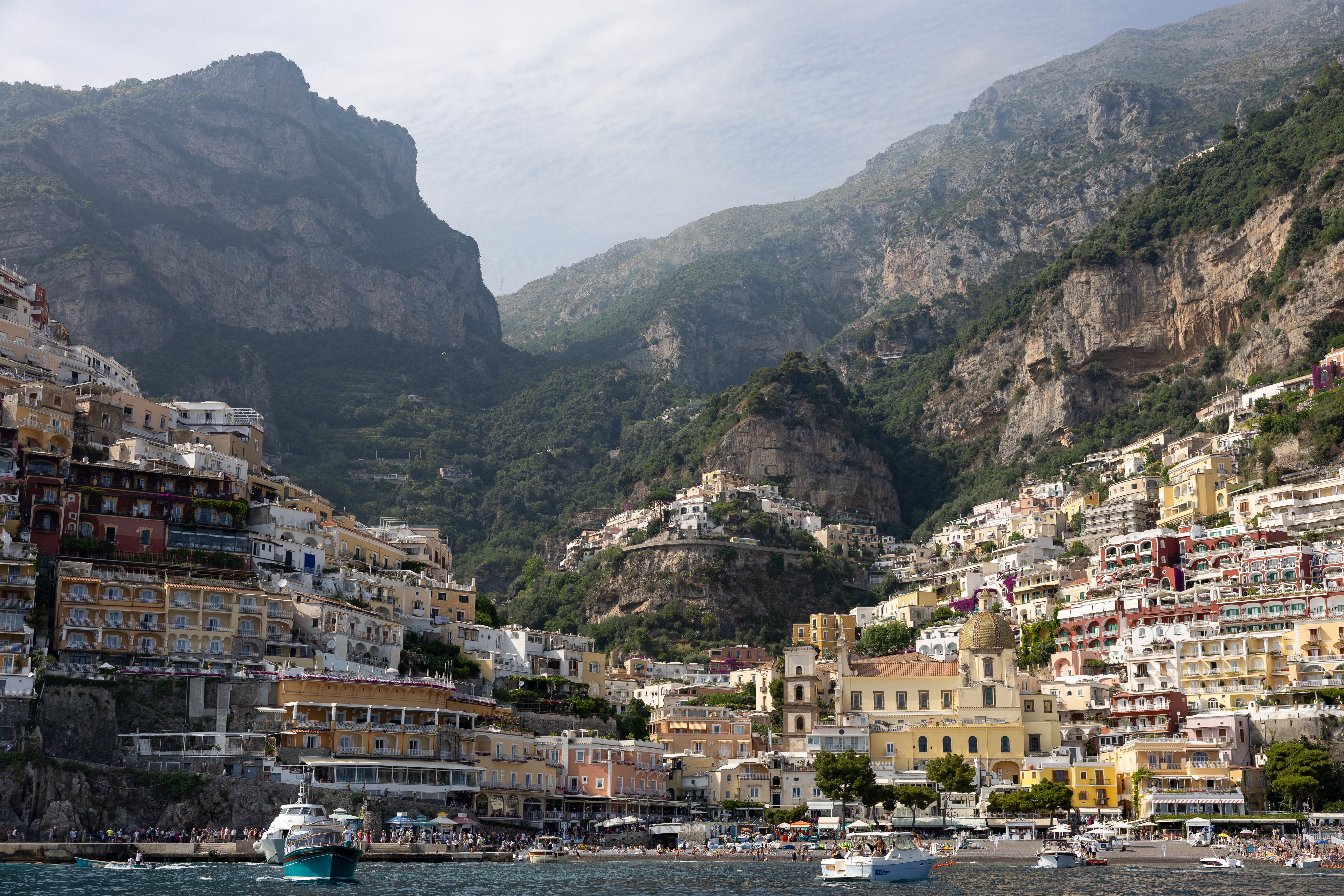 Wide-angle view of Positano’s colorful cliffside buildings and Church of Santa Maria Assunta as seen from the sea, backed by dramatic mountain peaks.