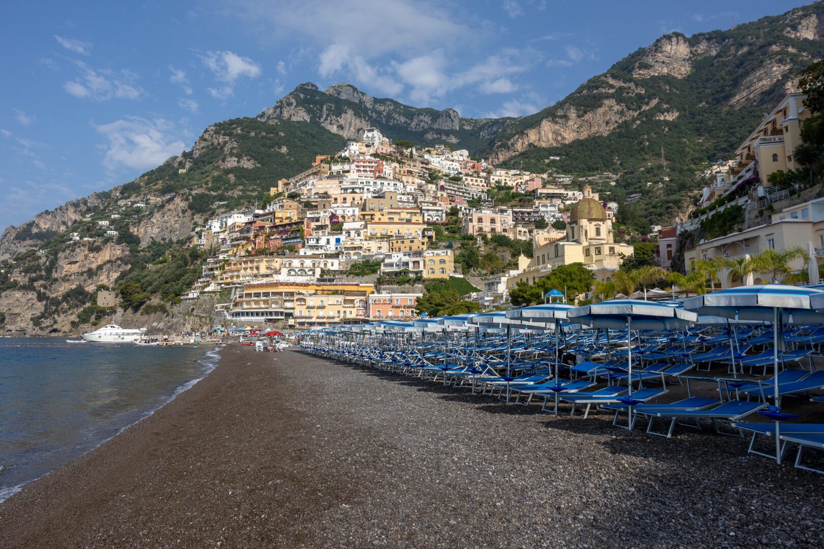 Rows of blue beach chairs and umbrellas line Positano’s Spiaggia Grande, with the town’s pastel buildings and iconic church dome rising against a backdrop of dramatic cliffs.