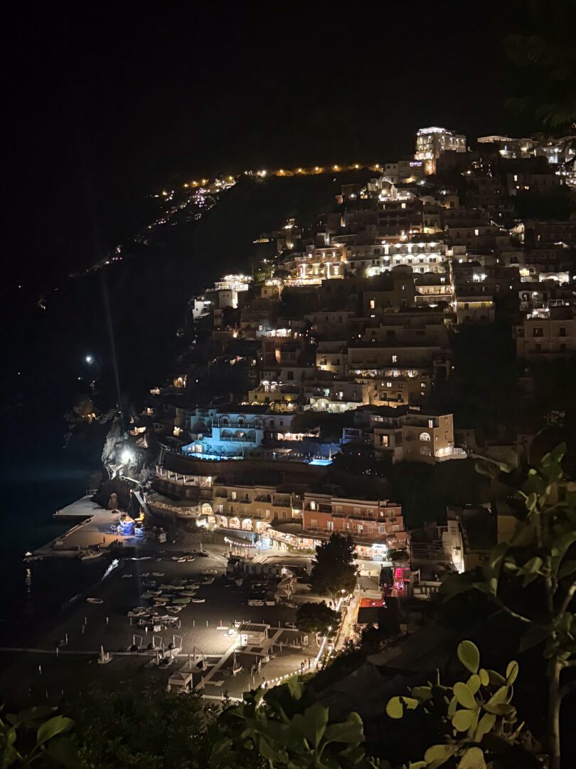 Positano illuminated at night, with hillside homes glowing above the dark shoreline and boats resting in the quiet marina.