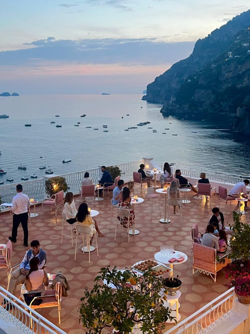 Guests sip cocktails on the elegant terrace of RED Cocktail and Champagne Bar in Positano at sunset, overlooking calm seas dotted with boats and the Amalfi Coast’s dramatic cliffs.