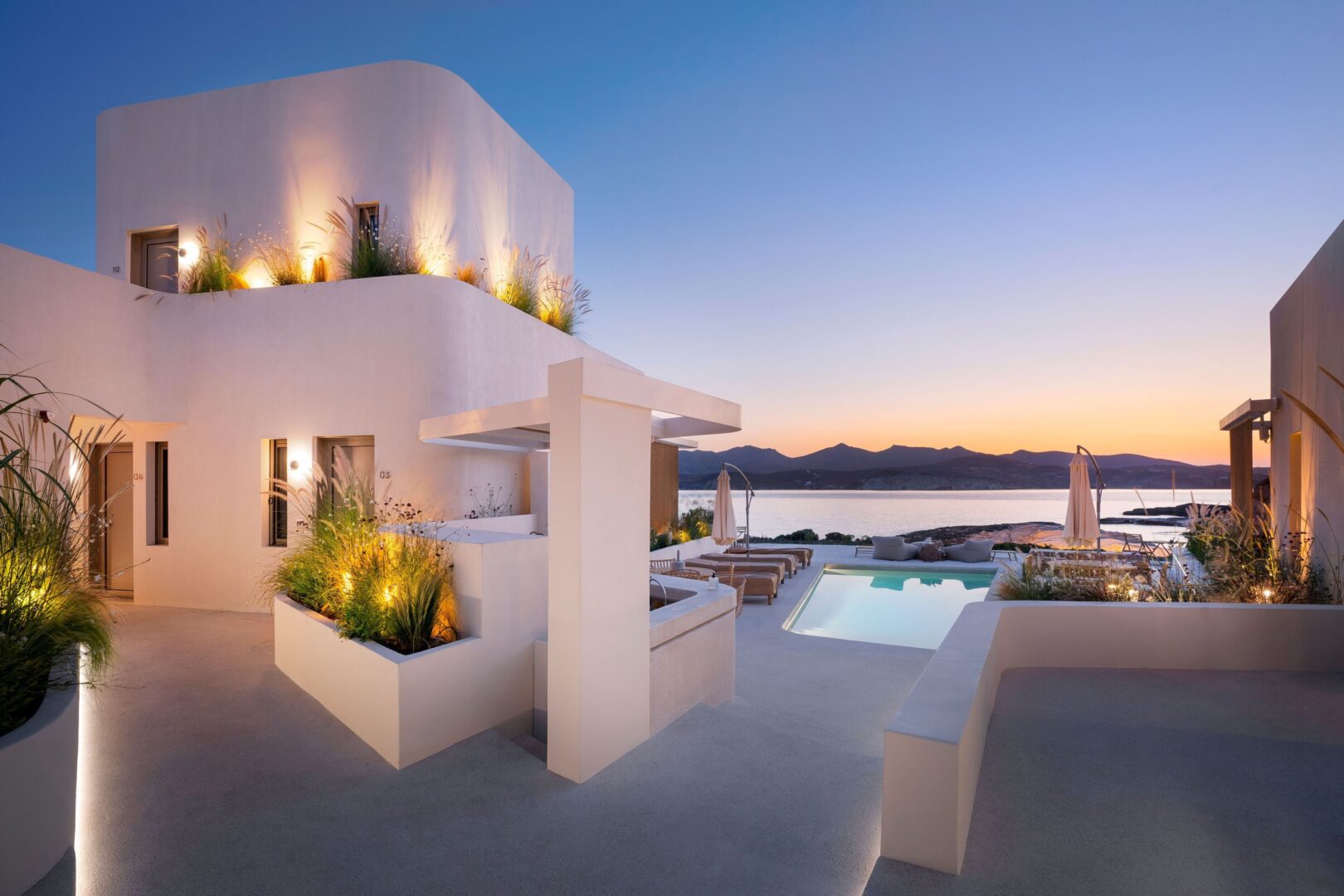 White Pebble Suites Milos at dusk with glowing modern architecture, a serene infinity pool, and a panoramic sea view near Pollonia.