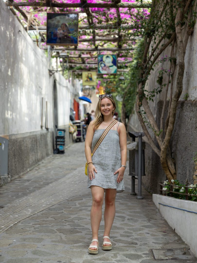 Woman poses under blooming bougainvillea and hanging artwork on the charming pedestrian street Via dei Mulini in Positano.