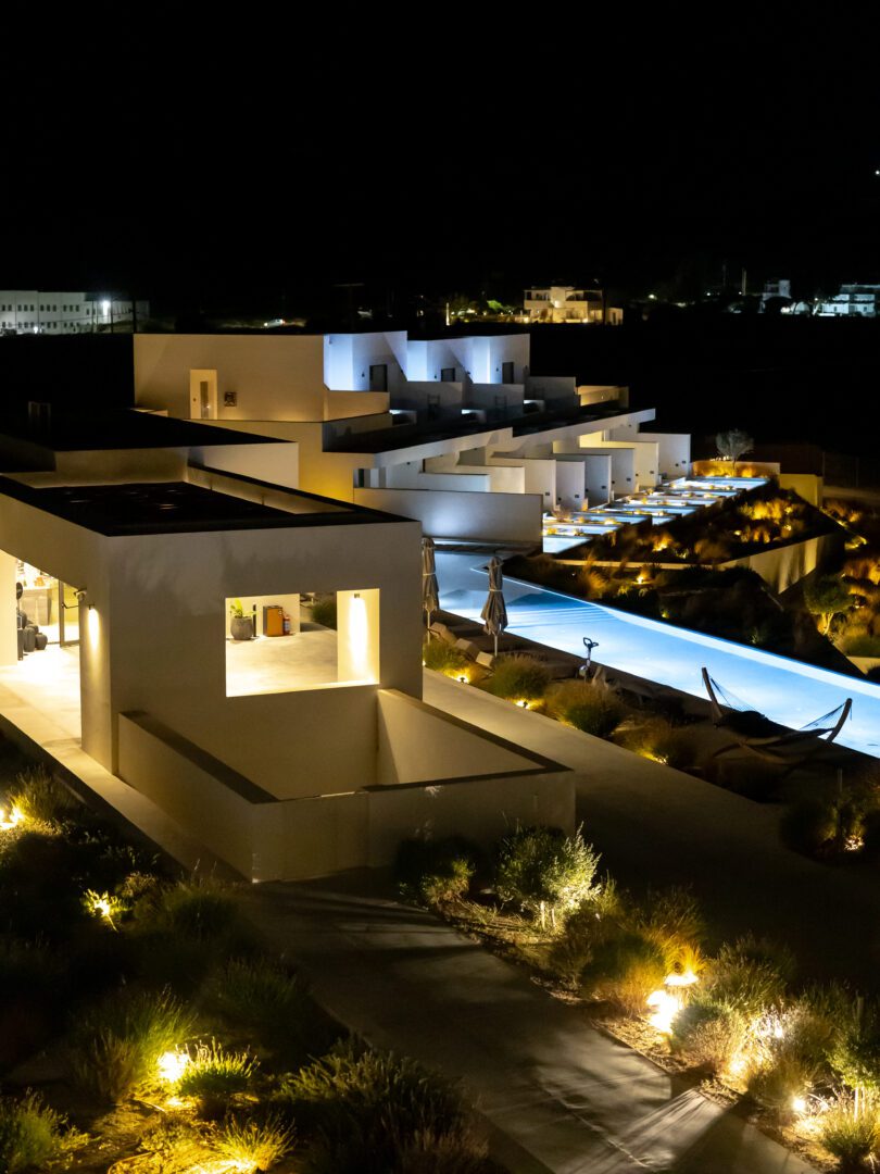 Illuminated Domes White Coast Milos exterior at night, showcasing modern Cycladic architecture and private plunge pools glowing against the dark sky.