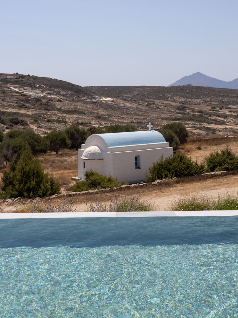 Sparkling infinity pool at Domes White Coast Milos overlooking a small whitewashed chapel with blue dome set against the rugged island landscape.