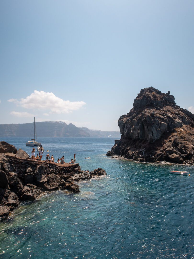 People gathered at Ammoudi Bay’s cliff-jumping spot in Santorini, with turquoise water, rocky cliffs, and a sailing boat in the distance.