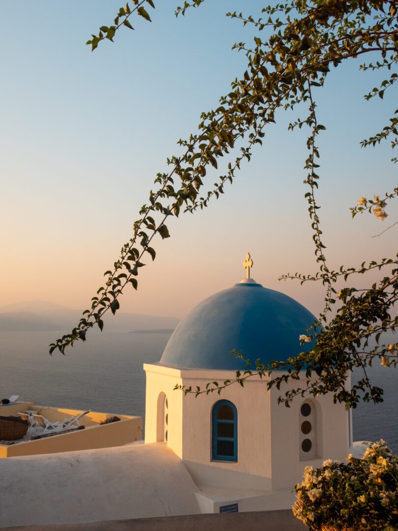 Blue-domed church in Oia, Santorini, glowing in the sunrise light with bougainvillea framing the view of the Aegean.
