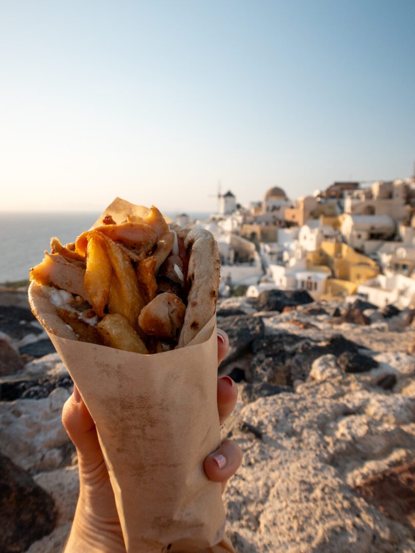 Chicken gyro from Pitogyros Traditional Grill House held in front of Oia Castle’s whitewashed buildings and windmill at sunset in Santorini.