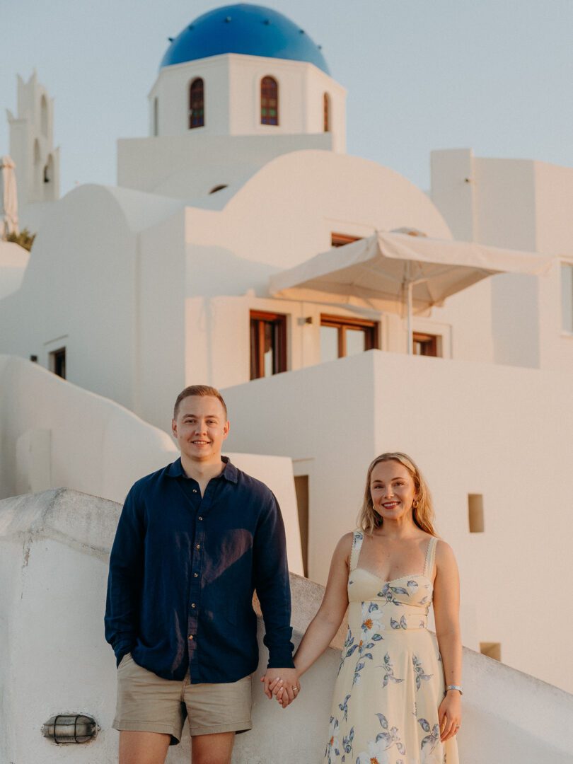 Couple holding hands in front of a blue-domed church and whitewashed architecture in Oia, Santorini at golden hour.
