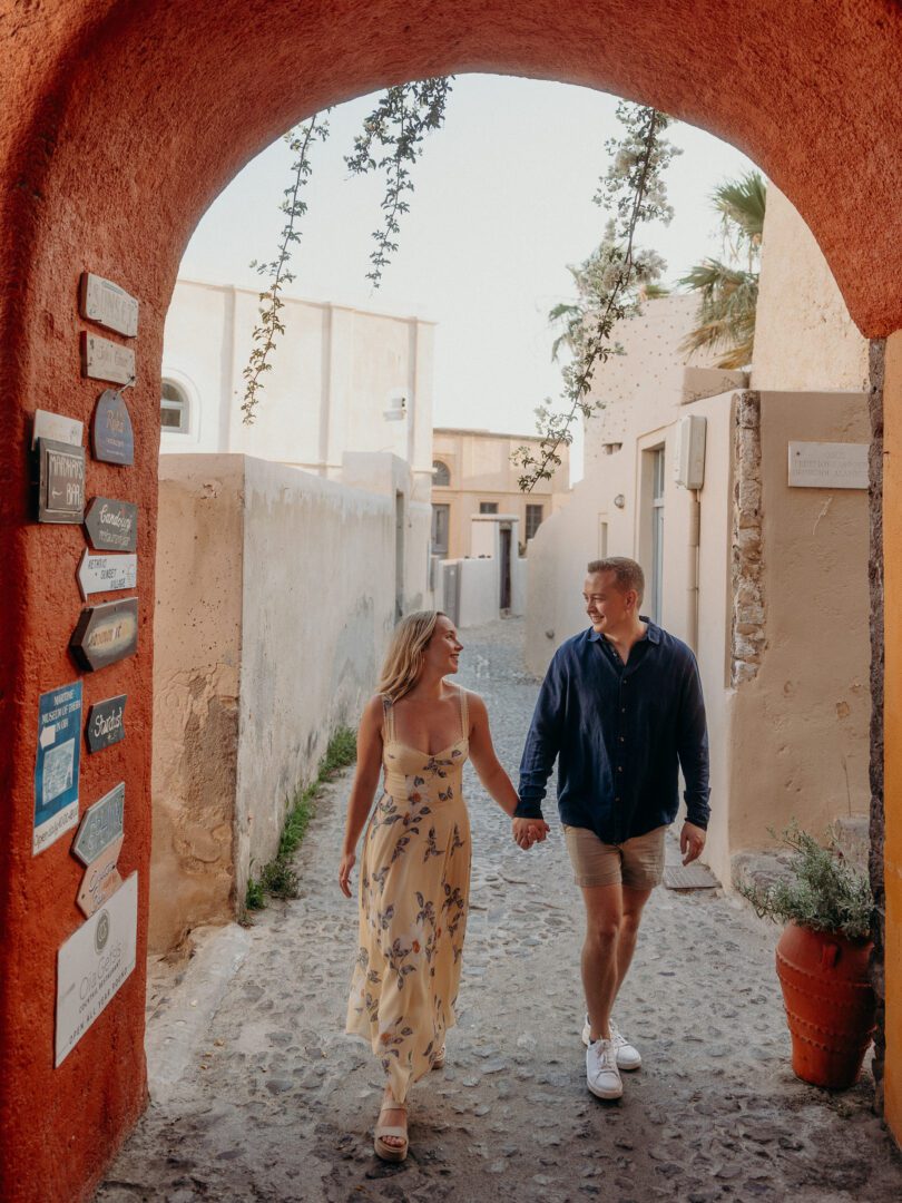 Couple walking hand in hand under a rustic orange archway in Oia’s charming alleys, during their Santorini Greece vacation.