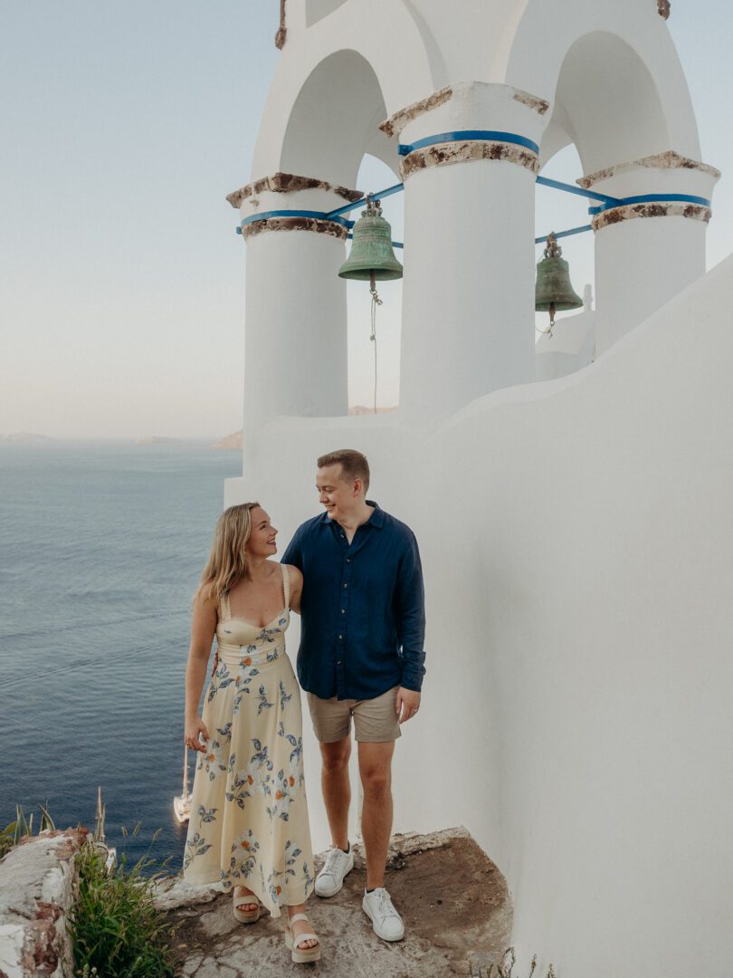 Couple smiling and walking beneath a whitewashed bell tower overlooking the Aegean Sea in Santorini.