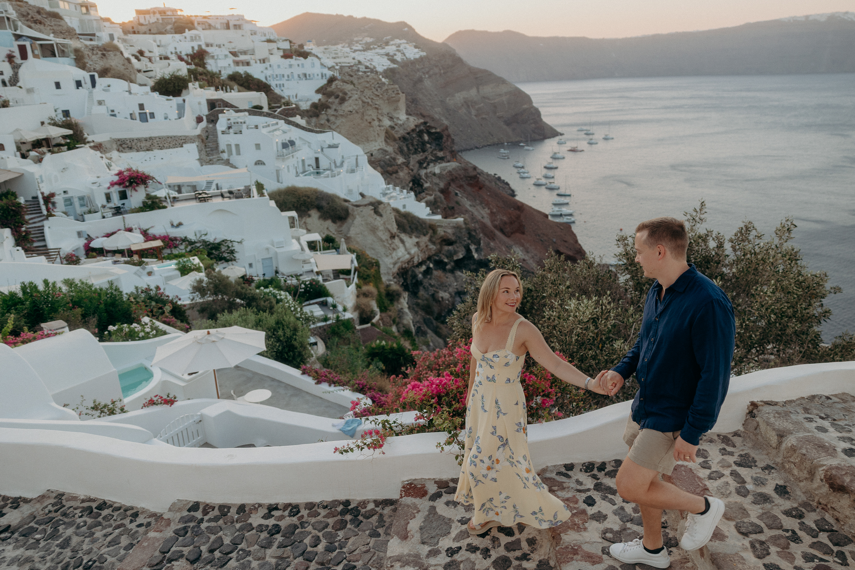 Couple walking hand in hand down stone steps in Oia, Santorini, with whitewashed houses, bougainvillea, and caldera views at sunrise.