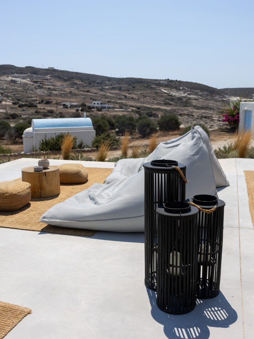 Relaxed seating with beanbags and lanterns at Domes White Coast Milos overlooking a whitewashed chapel and hillside views.