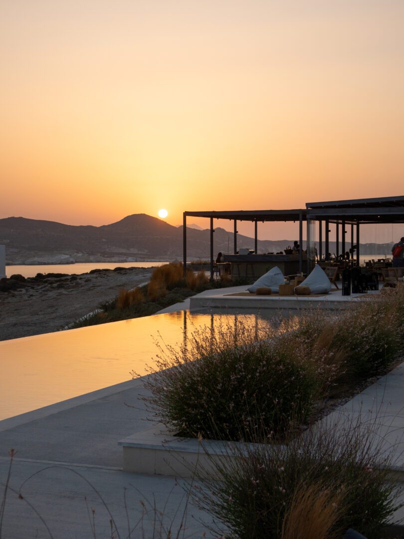 Golden sunset reflecting on the infinity pool at Domes White Coast Milos with views of the bar and distant hills.