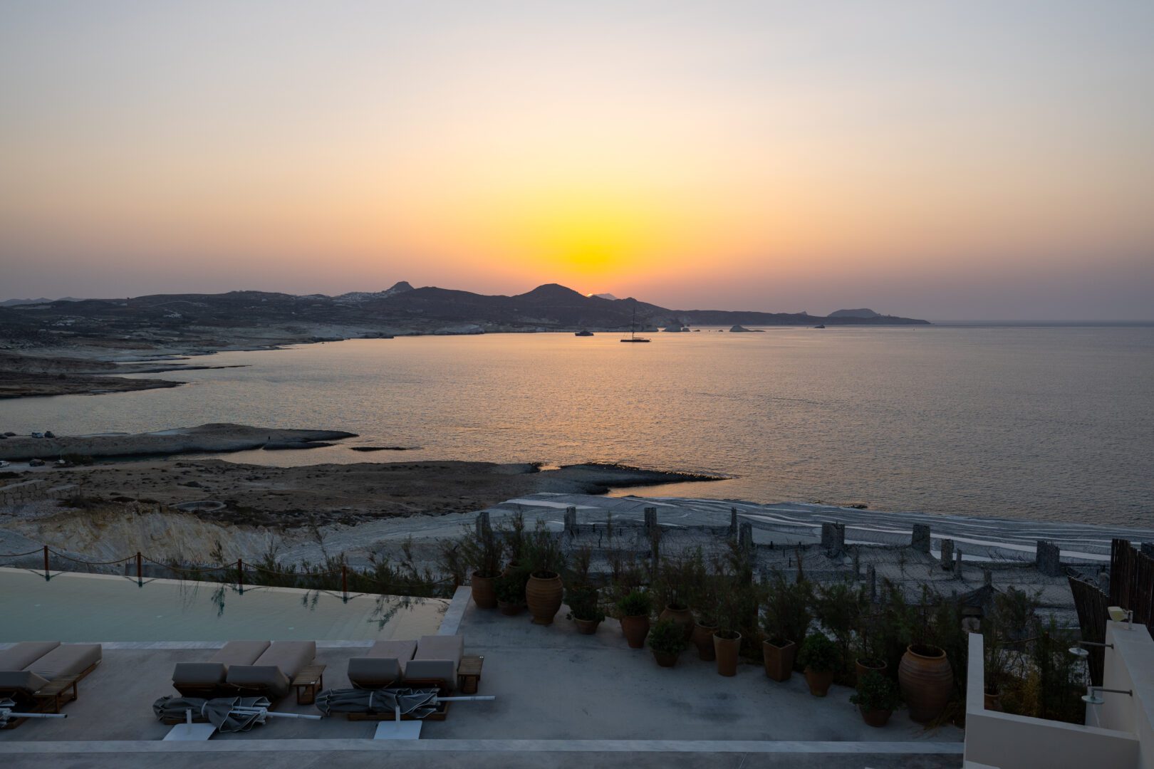 Stunning sunset over the Aegean Sea as seen from the terrace at Domes White Coast Milos with infinity pool loungers in the foreground.