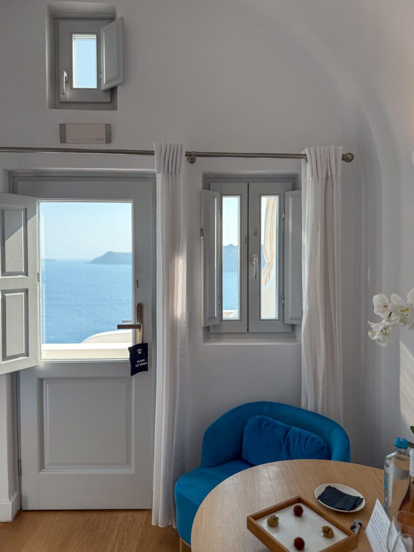 Junior suite interior at Katikies Hotel in Santorini with blue armchair, round table, and caldera views through the windows.