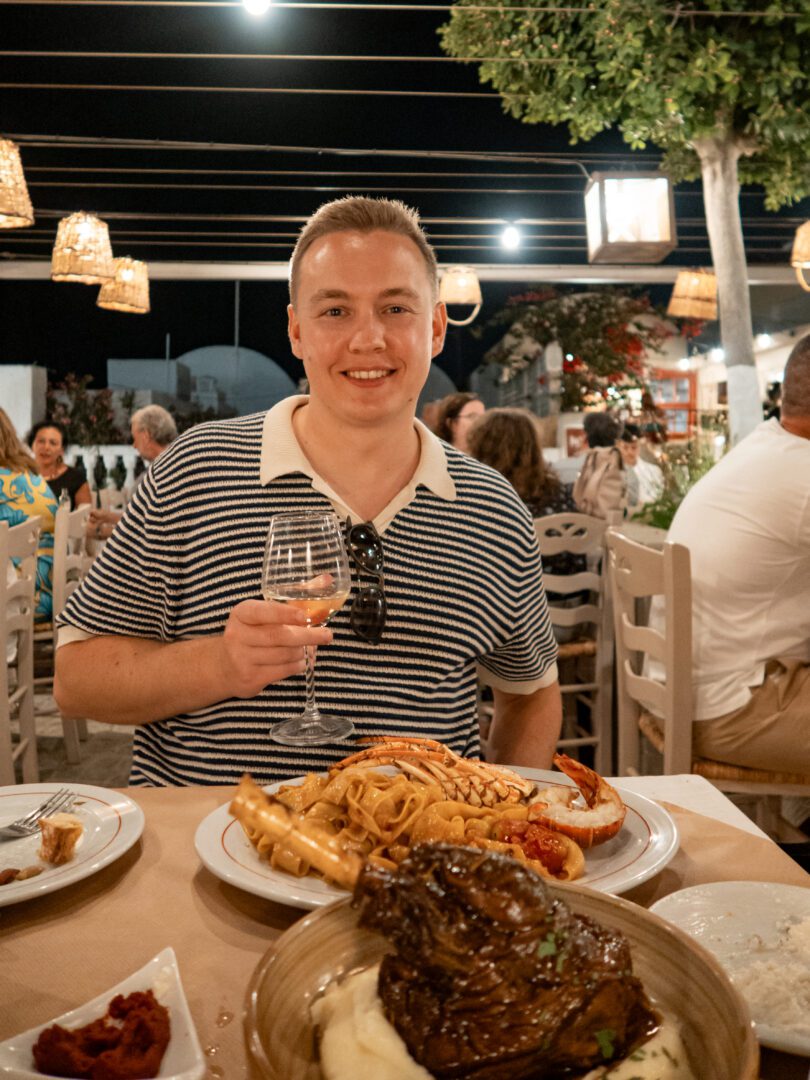 Man enjoying a glass of white wine with lamb shank and lobster pasta at Fino Taverna in Oia, Santorini.