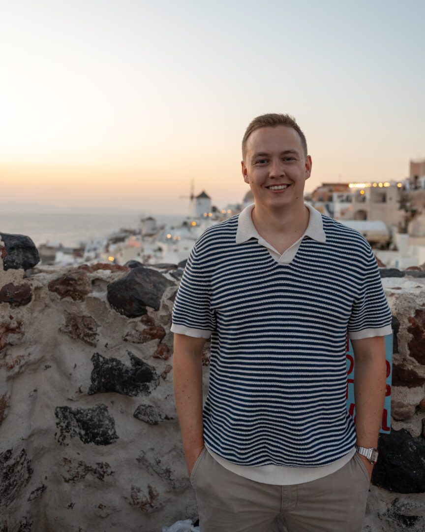Man smiling at Oia Castle at sunset during a Santorini Greece vacation, with the village’s whitewashed houses and windmill in the background.