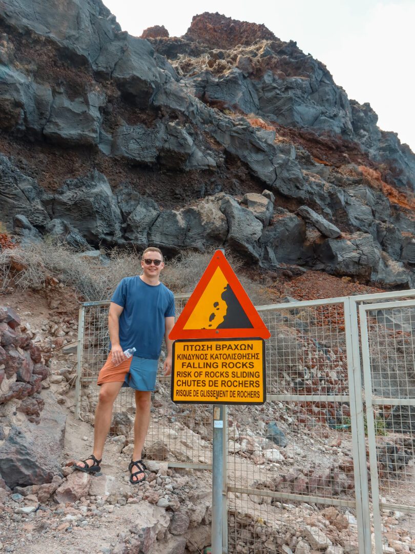 Man standing beside a warning sign for falling rocks near the gate to Ammoudi Bay in Santorini, Greece.