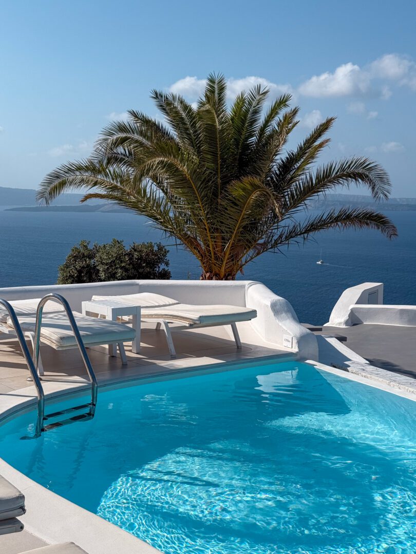 Lounge chairs by the turquoise pool with palm tree and caldera views at Katikies Hotel in Santorini.