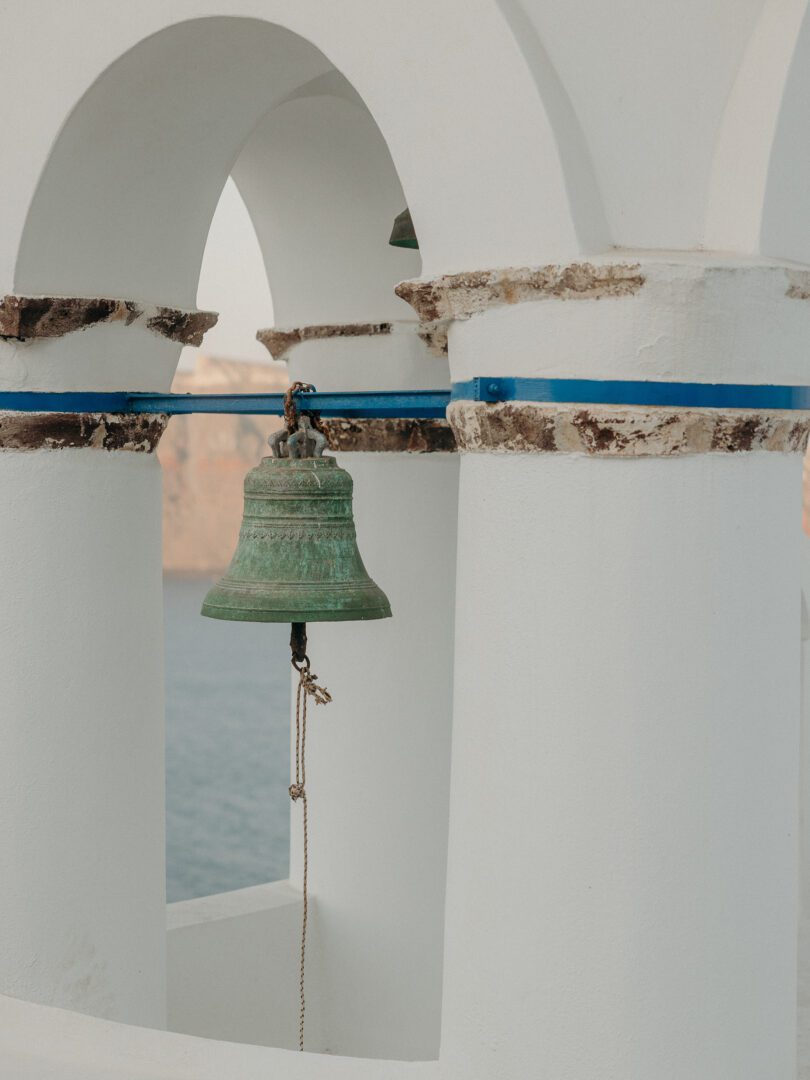 Close-up of a traditional green church bell hanging in a whitewashed bell tower in Santorini, Greece.