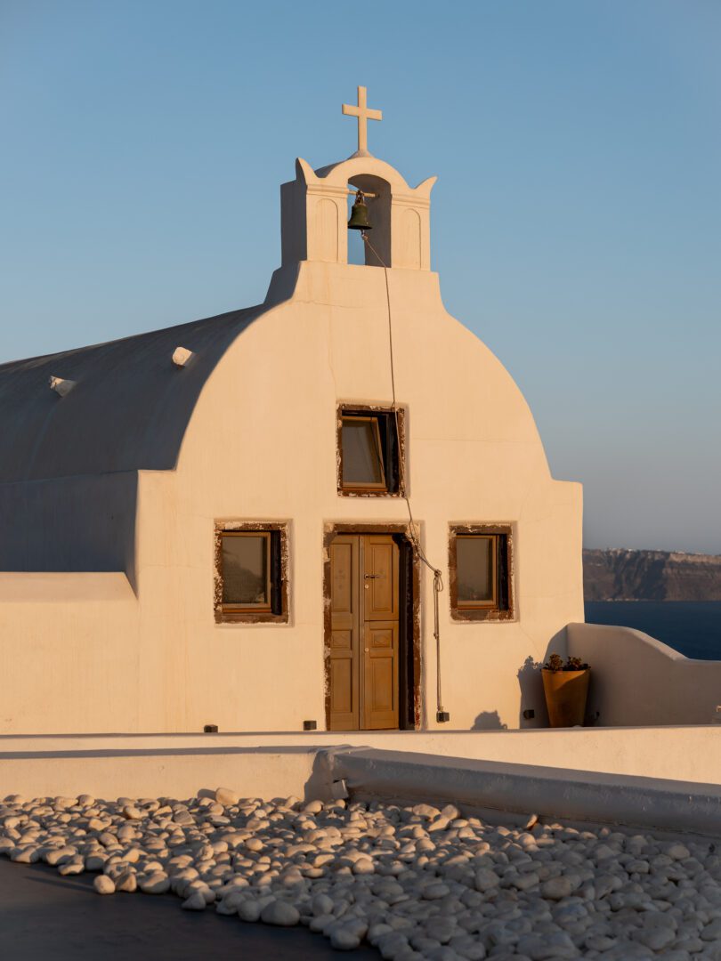 Traditional whitewashed church with a bell tower and cross in Oia, Santorini, overlooking the caldera at golden hour.