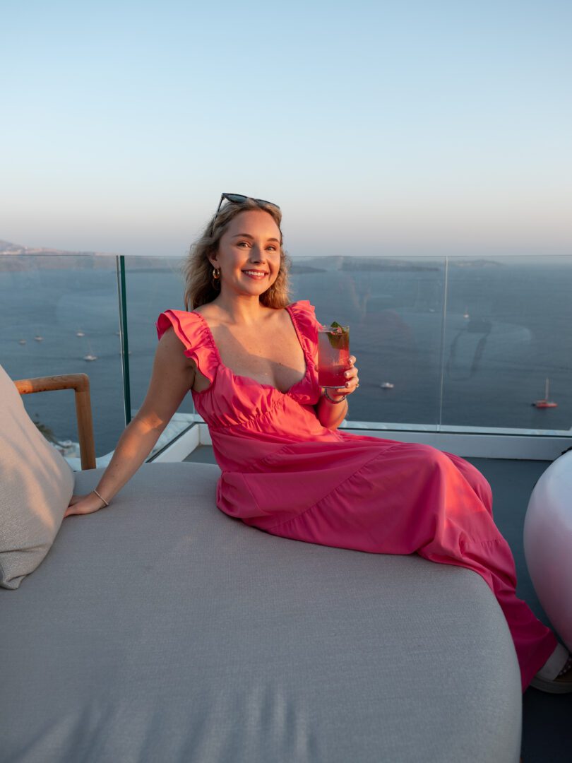 Woman in a pink dress enjoying a cocktail at Canaves Sunday Veranda Bar with sweeping caldera views at sunset during her Santorini Greece vacation.